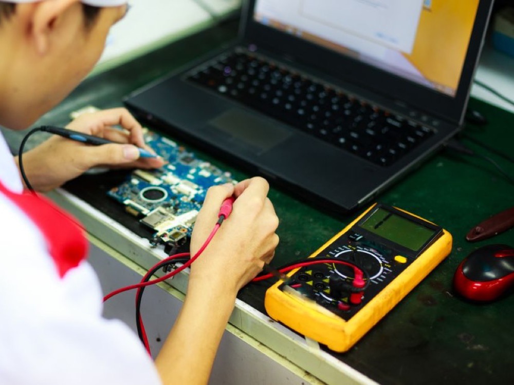 Técnico utiliza pontas de prova de um multímetro para verificar uma placa eletrônica ao lado de um notebook em bancada.