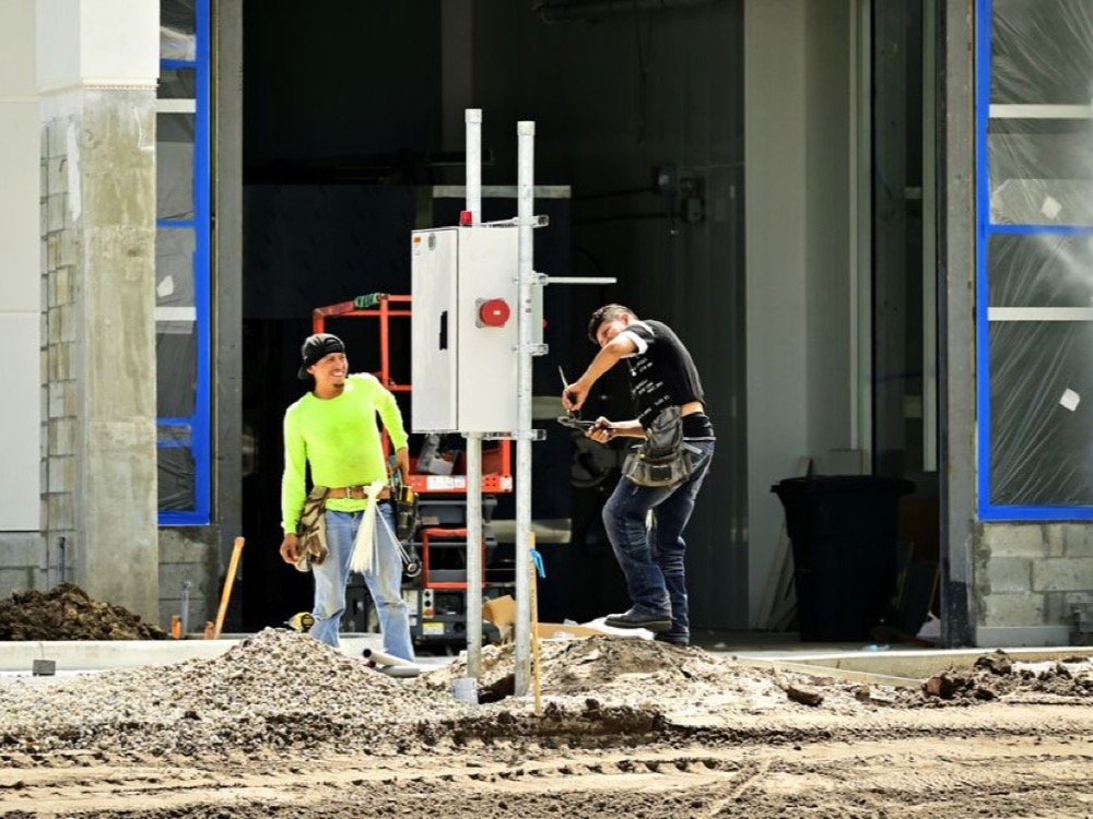 Dois trabalhadores realizam serviços em uma obra, próximo a um quadro elétrico instalado sobre uma estrutura metálica.
