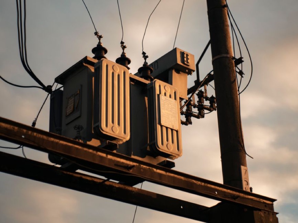 Transformadores elétricos instalados em um poste com cabos e conexões da rede de distribuição de energia ao entardecer. Tudo para explicar qual o melhor transformador de 110 para 220.