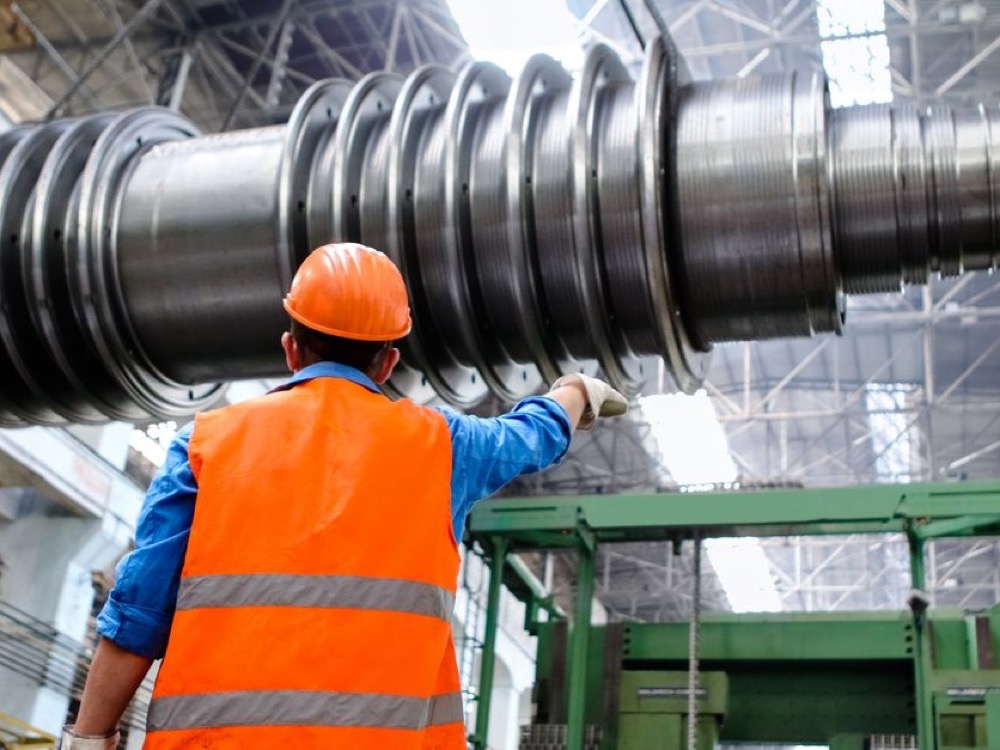 Trabalhador com capacete e colete de segurança observa um grande equipamento metálico instalado no interior de uma fábrica industrial.