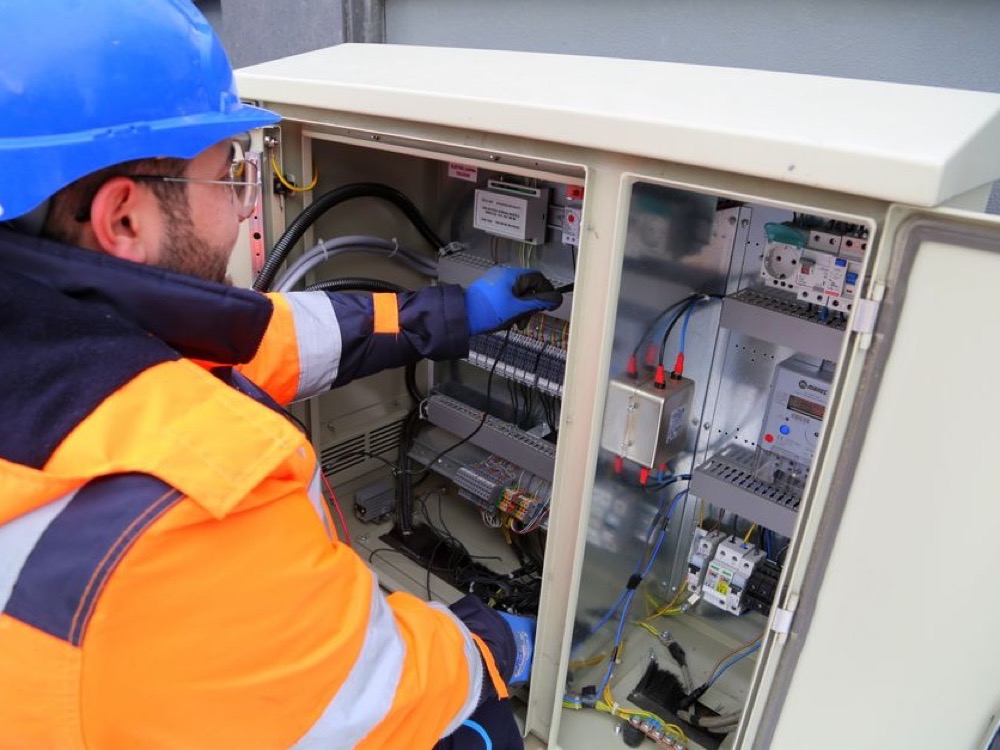 Eletricista com capacete azul e uniforme de proteção verifica os componentes internos de um painel elétrico industrial aberto.